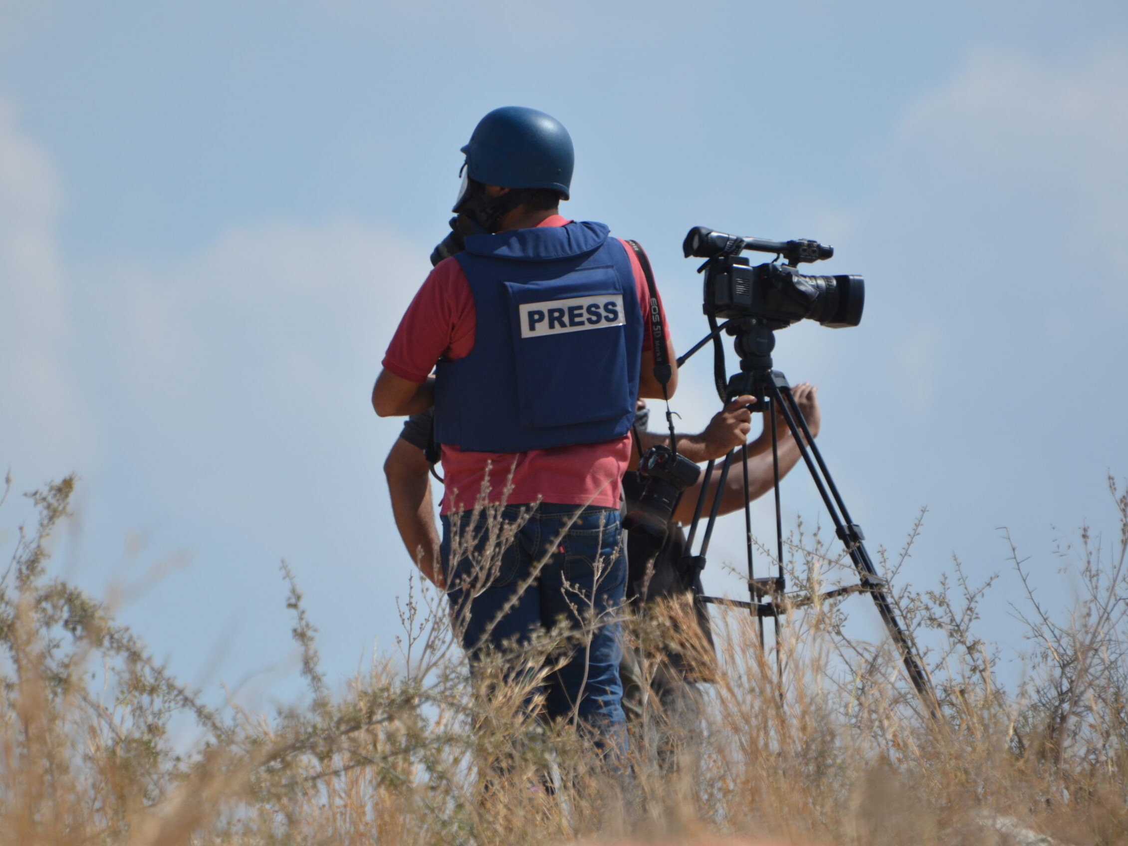 A journalist and a cameraperson wearing protective gear stand in a field.
