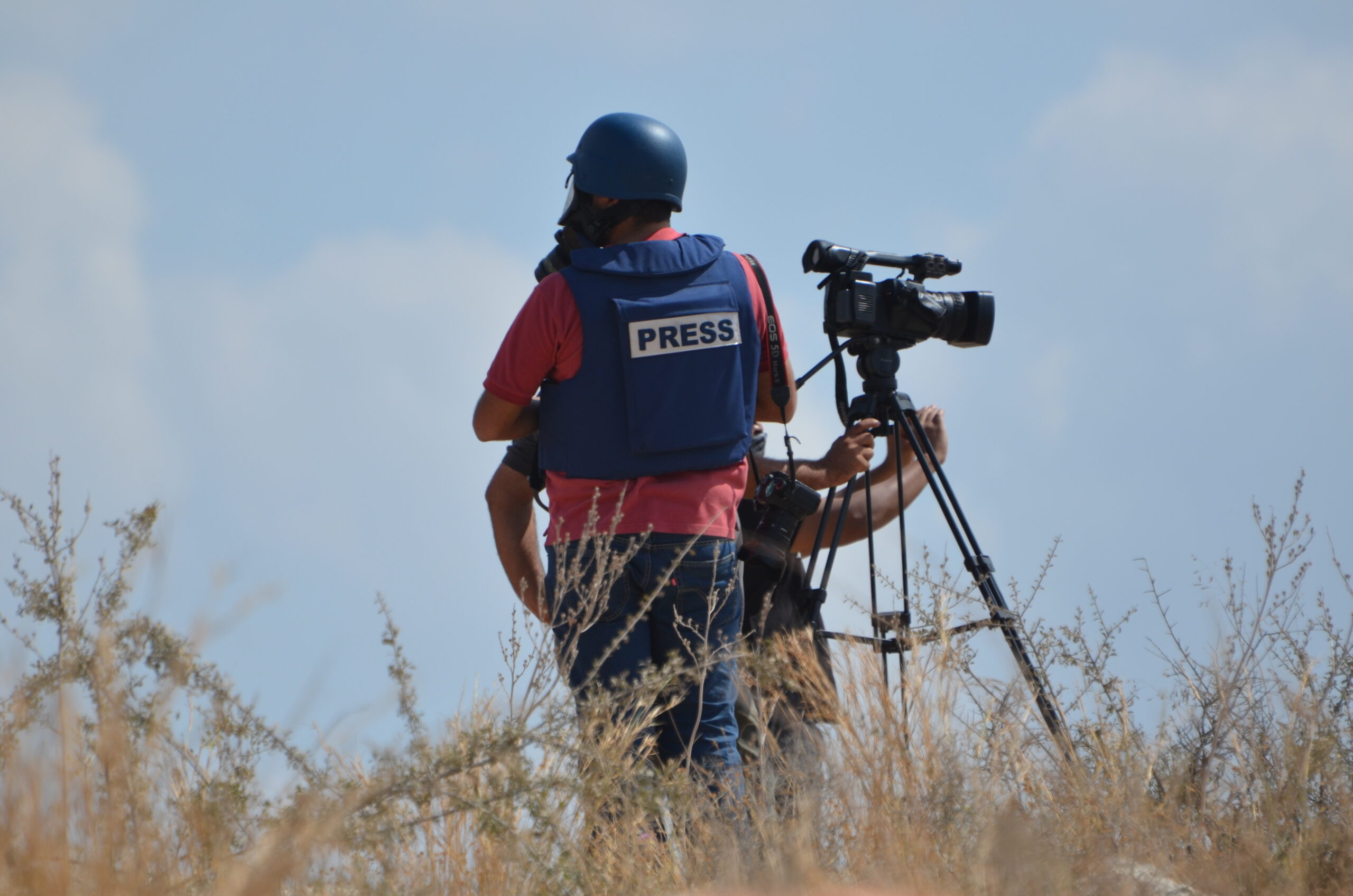A journalist and a cameraperson wearing protective gear stand in a field.