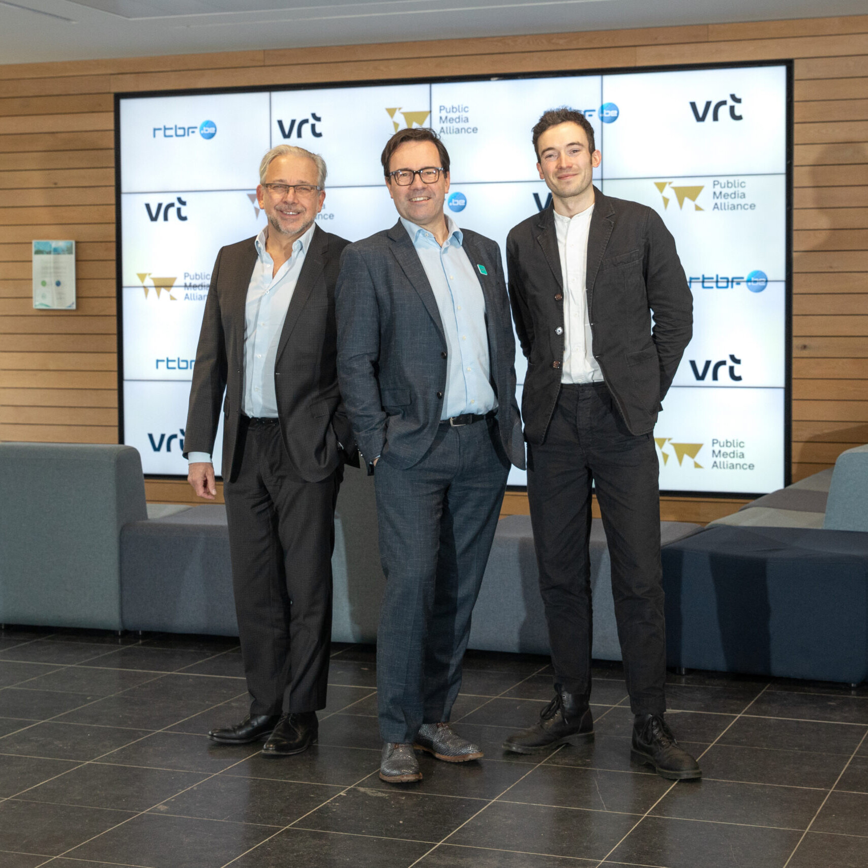 Three men stand in front of a screen with the VRT, PMA, and RTBF logos on it.