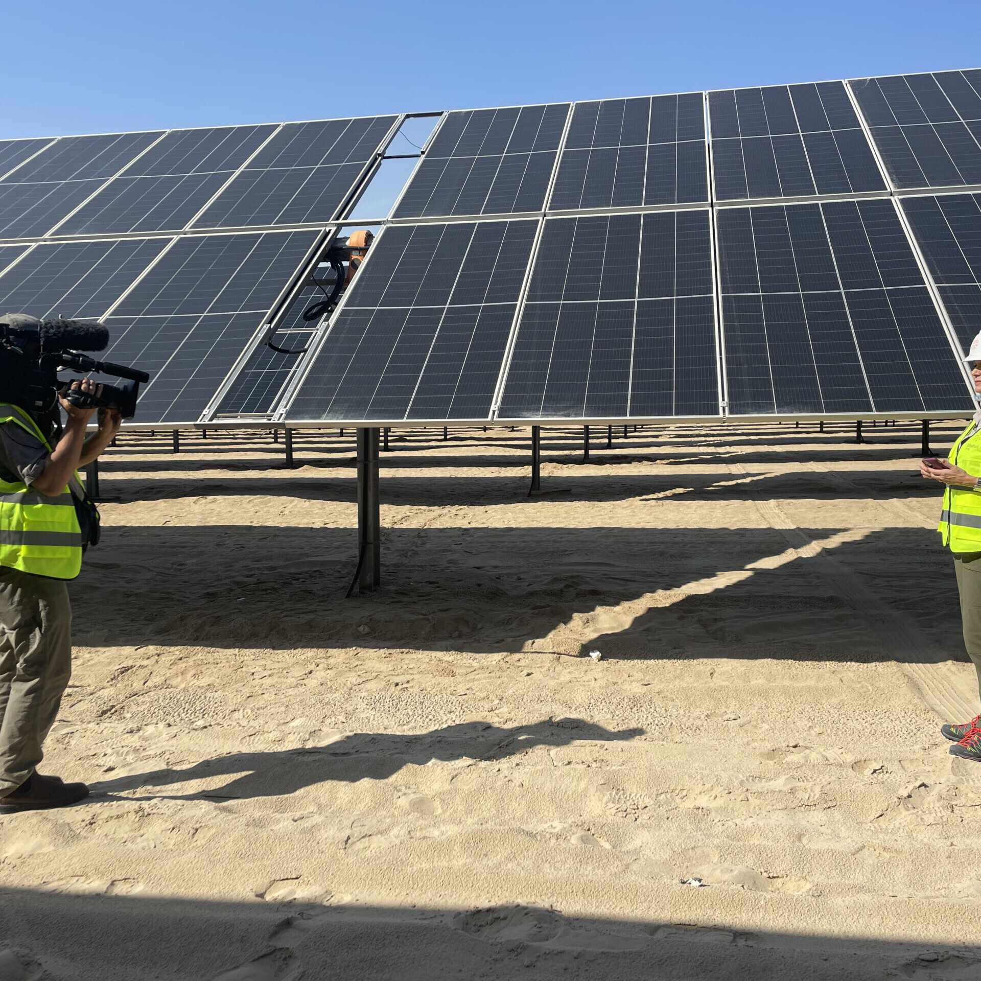 A cameraperson and a reporter stand in front of an enormous solar panel.