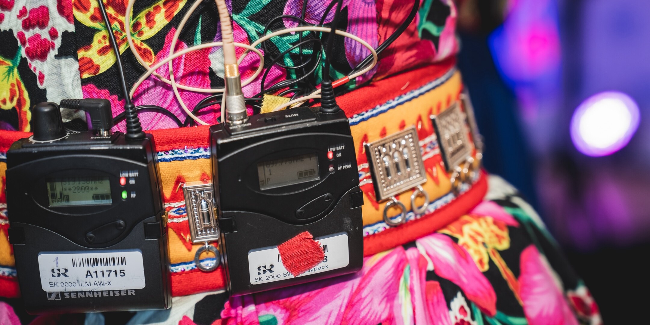 A closeup image of a colourful dress with two radio microphone sets and wires attached to the belt.