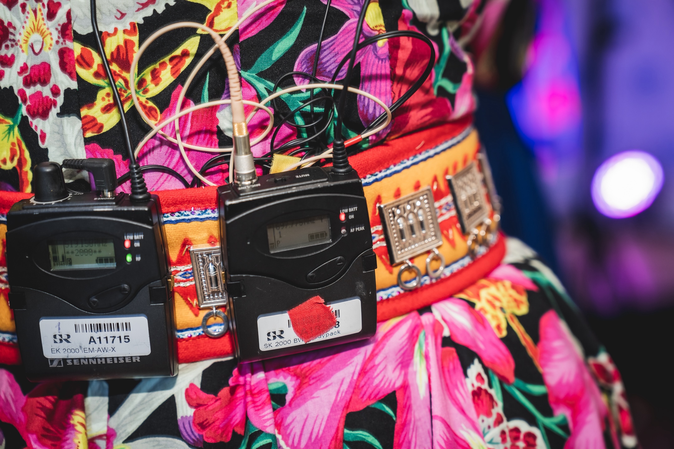 A closeup image of a colourful dress with two radio microphone sets and wires attached to the belt.
