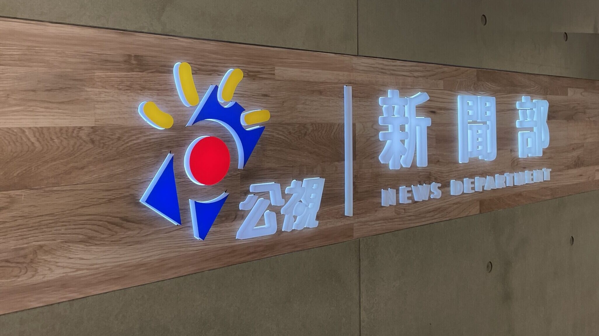 A wooden board runs horizontally along a concrete wall. On the wooden board is a colour logo for PTS, and a sign which reads 'News Department'.