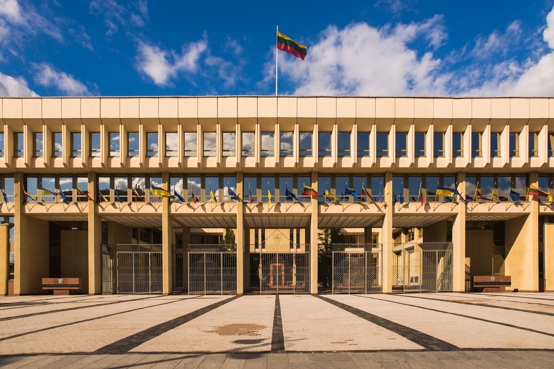 A rectangular concrete building bathed in sun, with the Lithuanian flag on a flagpole.