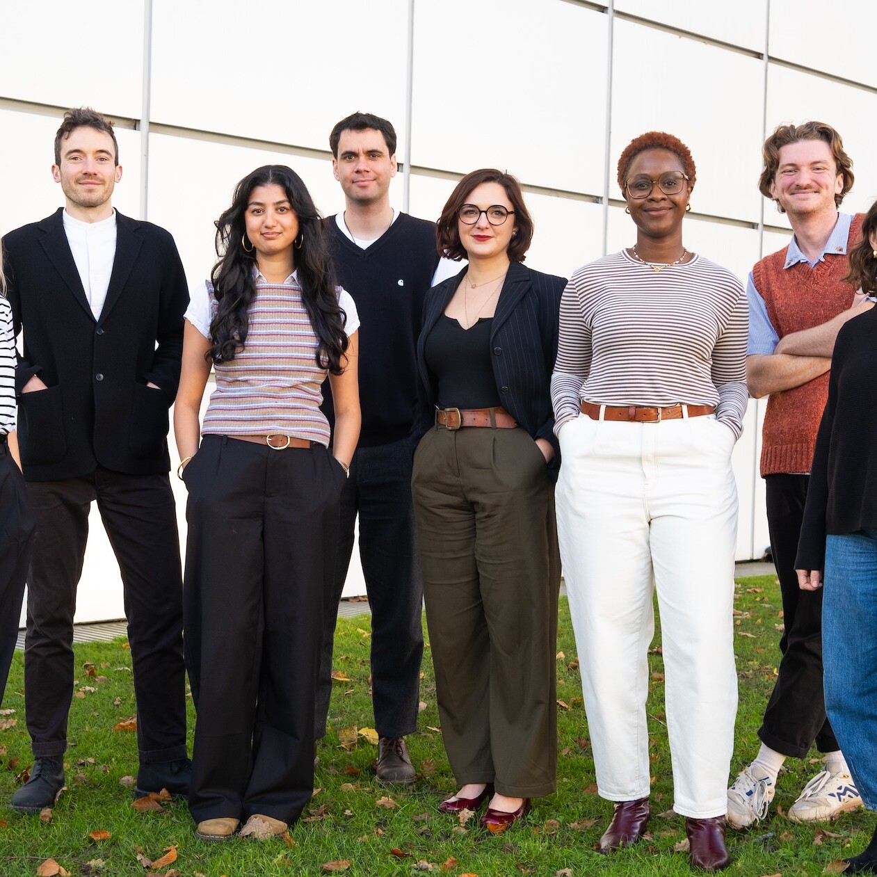 Eight people standing on green grass with a big white wall behind