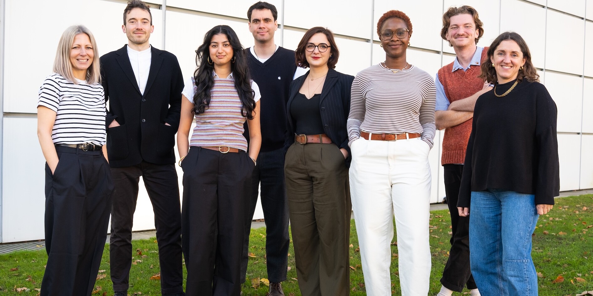 Eight people standing on green grass with a big white wall behind