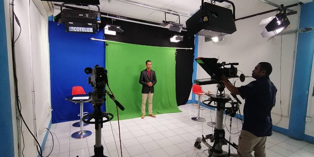 A wide angle of a TV studio with one person operating a camera, and a presenter standing in front of a green screen.