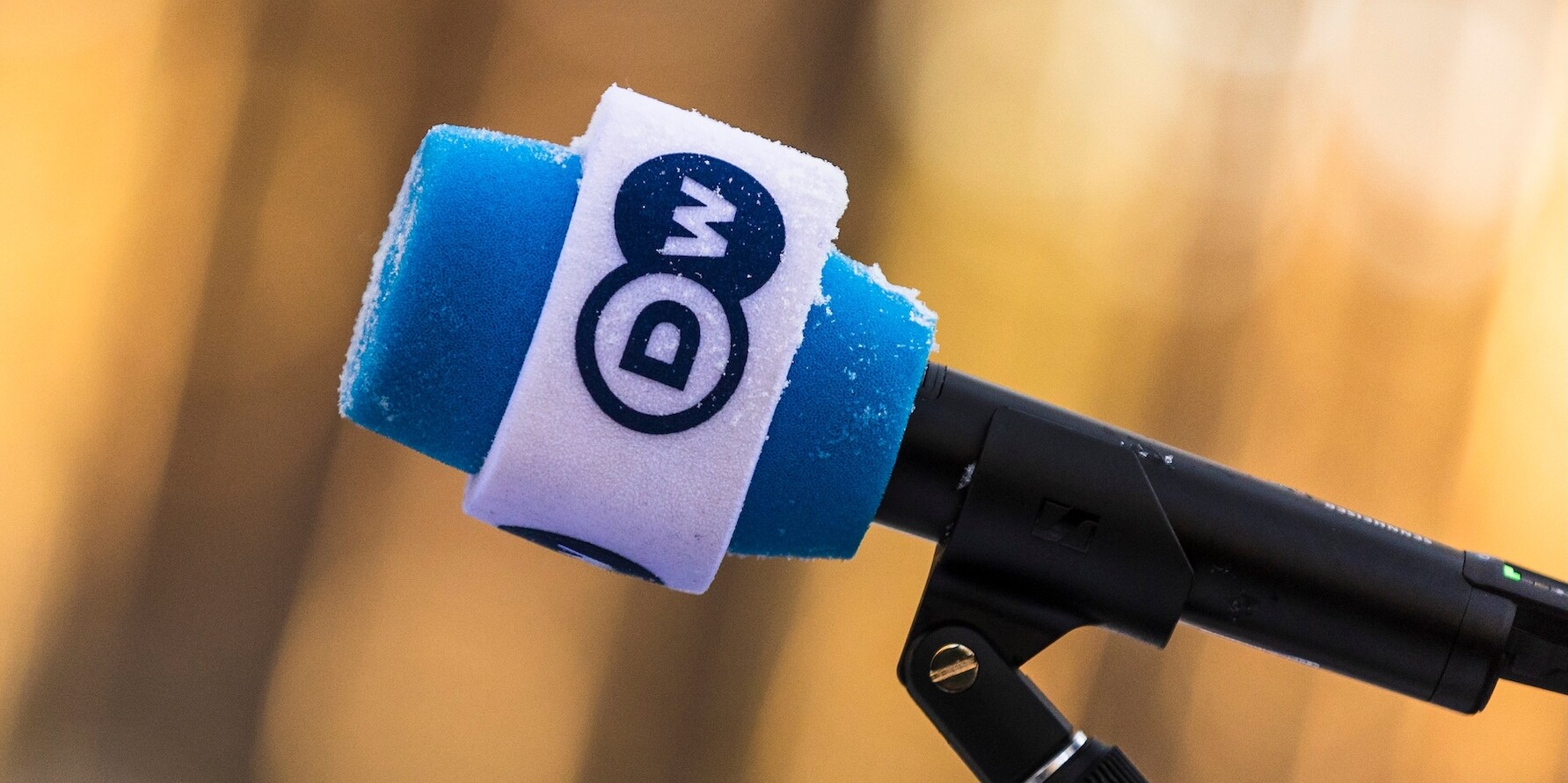 A closeup of a microphone on a holder. The microphone head is blue with a white band around it, with the DW logo on.