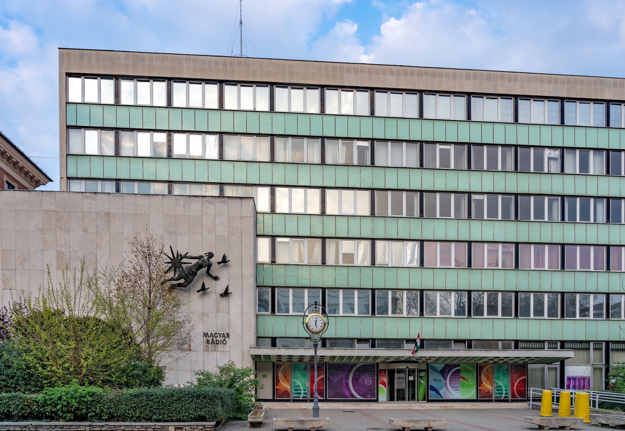 A 20th century office block made of concrete and windows. In front of the building is a concrete wall with a Magyar Radio logo.
