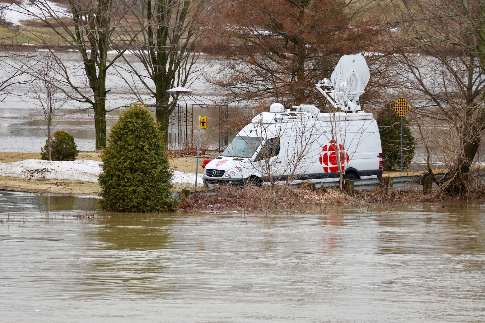 A white van with a satellite on top, and the CBC/Radio-Canada logo on the side is parked on the road, as it is surrounded by flooding on all sides.