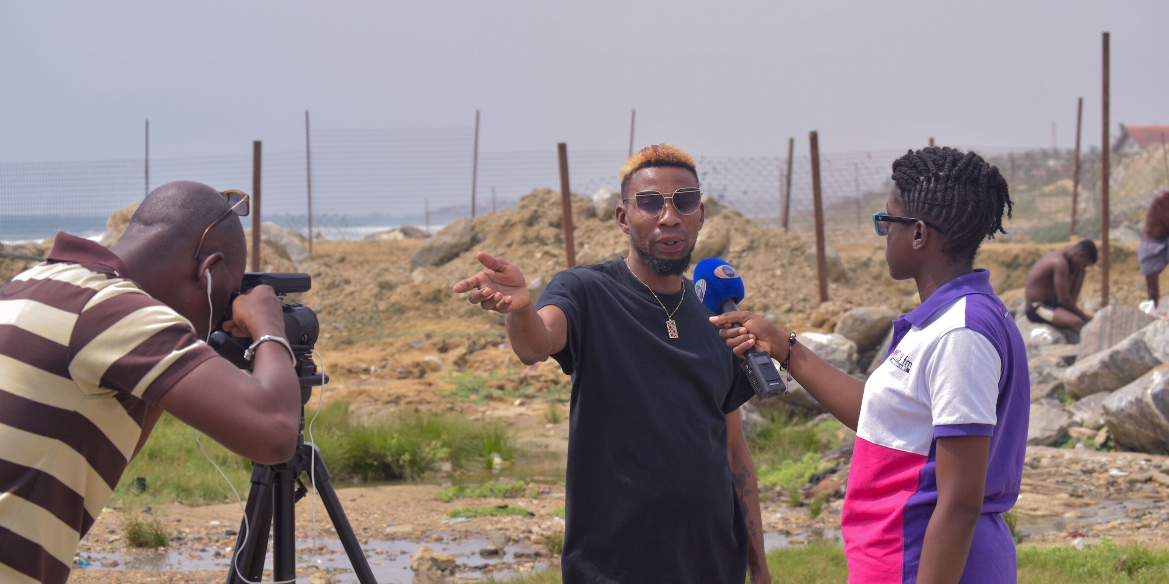 A Ghanaian woman journalist holds a microphone and is interviewing a man, who is gesturing with his left hand. A man films the interview with a camera on a tripod.
